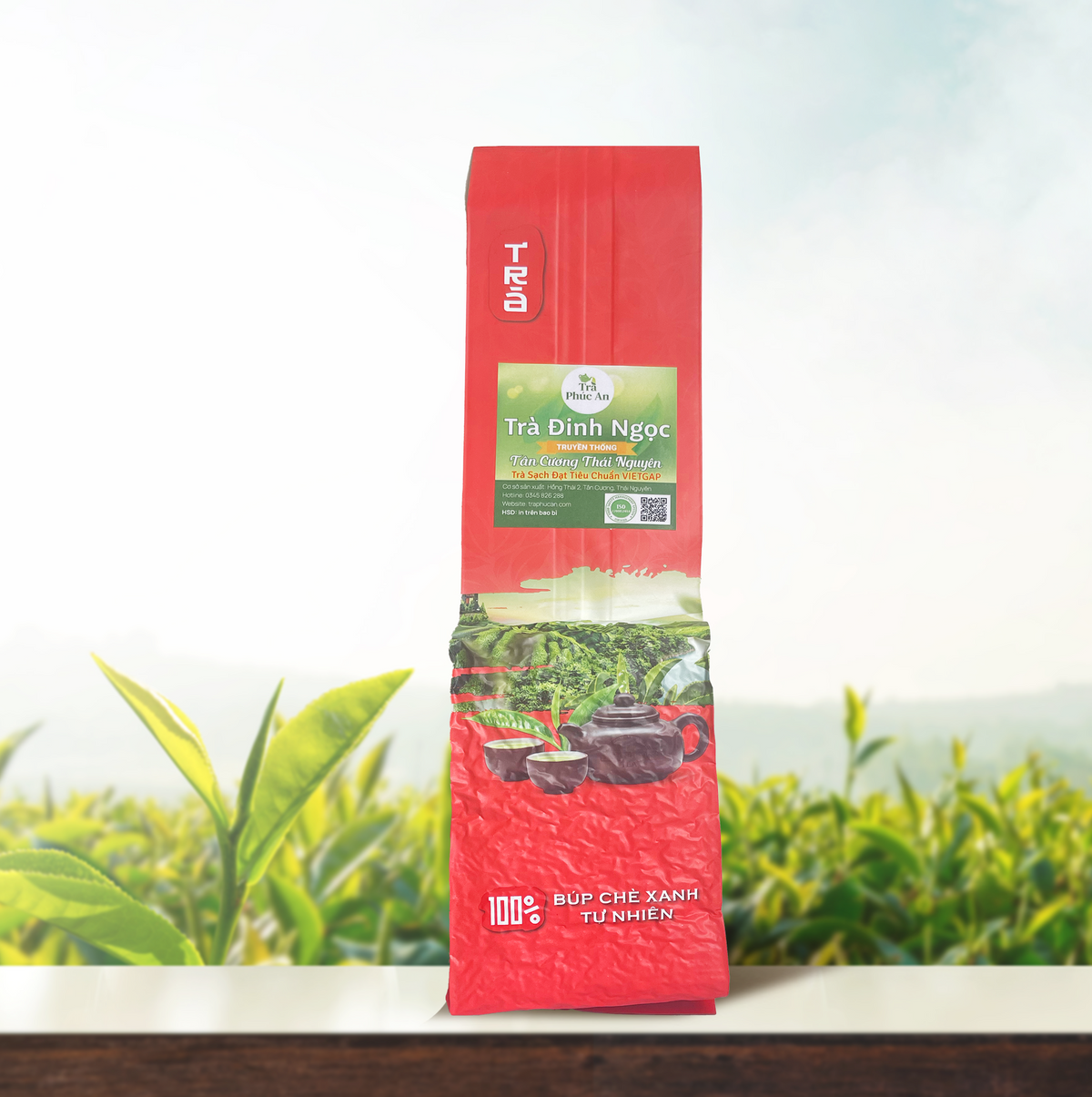 Gói Trà Đinh Ngọc nhãn Phúc An màu xanh trên bao bì đỏ, chụp chính diện trước nền đồi chè.