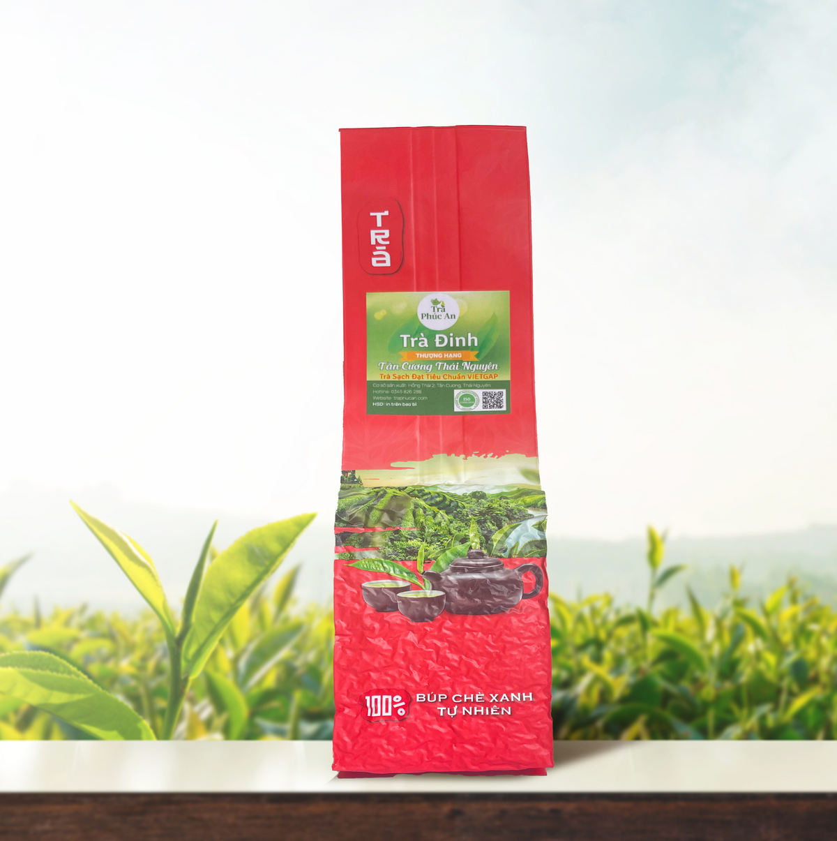 Bao bì Trà Đinh màu đỏ, nhãn xanh và hình ấm chén, chụp chính diện trên nền đồi chè.