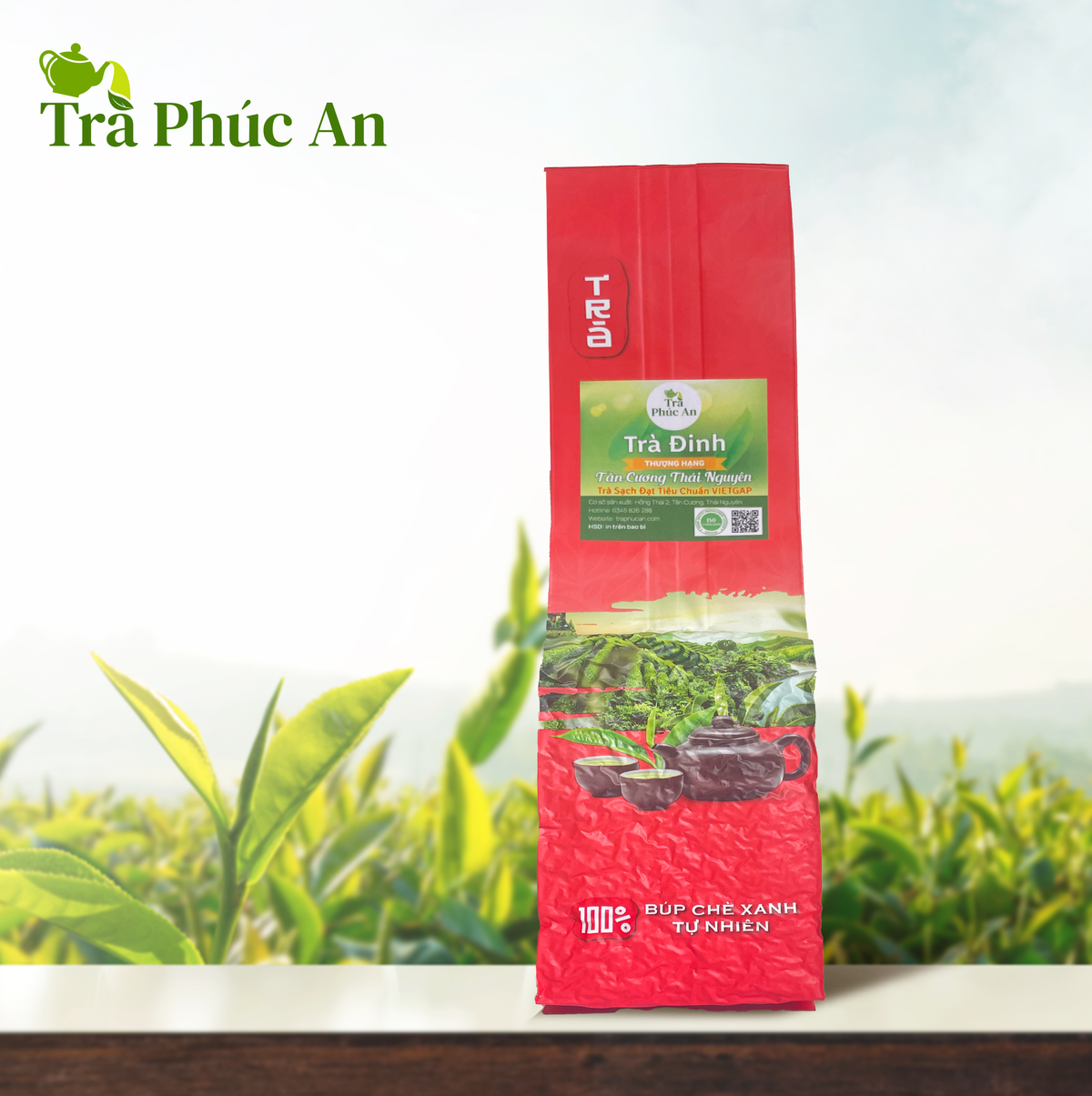 Gói Trà Đinh nhãn Phúc An màu xanh trên bao bì đỏ, đặt trước nền đồi chè có logo Trà Phúc An.