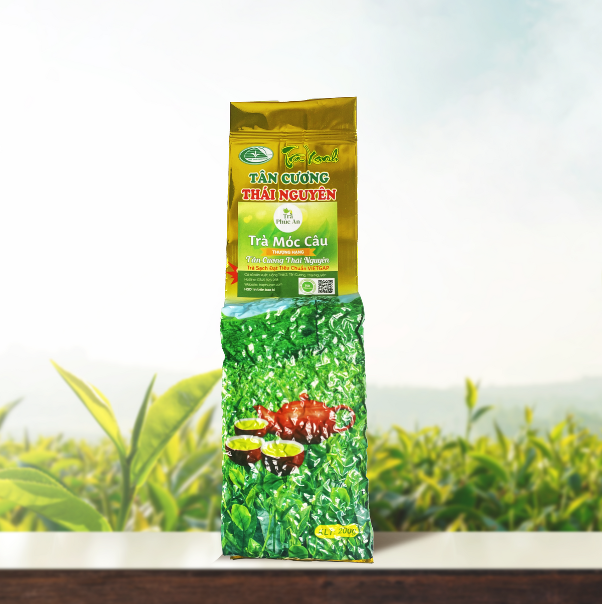 Gói Trà Móc Câu Thượng Hạng nhãn xanh, phần đầu màu vàng, đặt đứng trên nền đồi chè.