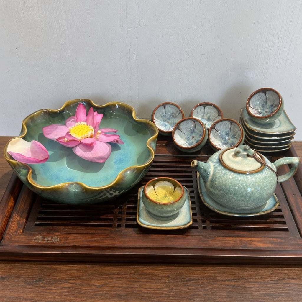 Bộ Trà Men Thỏ Ngọc màu xanh xám bày trên khay gỗ, cạnh bát thả hoa sen hồng; chén và đĩa được xếp chồng.