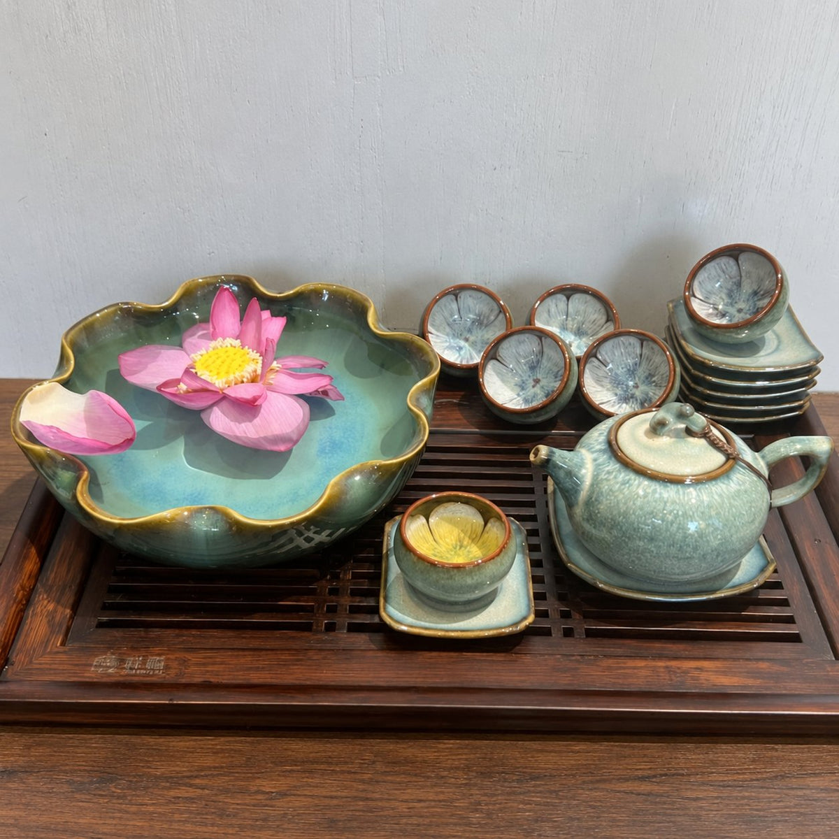 Bộ Trà Men Thỏ Ngọc màu xanh xám bày trên khay gỗ, cạnh bát thả hoa sen hồng; chén và đĩa được xếp chồng.